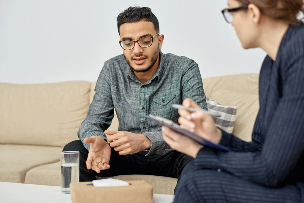 a man with glasses sits on a couch while his therapist takes notes and talks to him about alcohol addiction rehab expectations