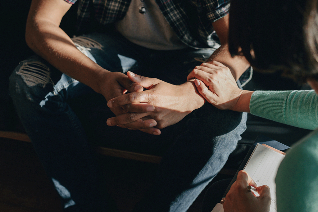 a person is sitting and has hands folded in their lap while someone else reaches out and touches their forearm explaining they will help them find a alcohol detox program in houston, tx